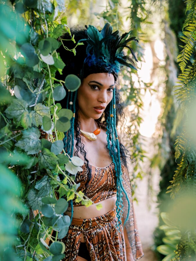 Performer com toucado de plumas azuis entre folhagem, no evento WILD