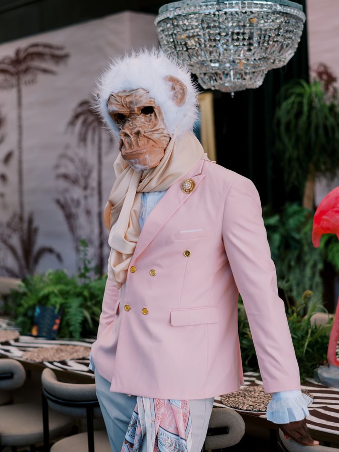 Performer com máscara de macaco e casaco cor de rosa, sob um lustre de cristais