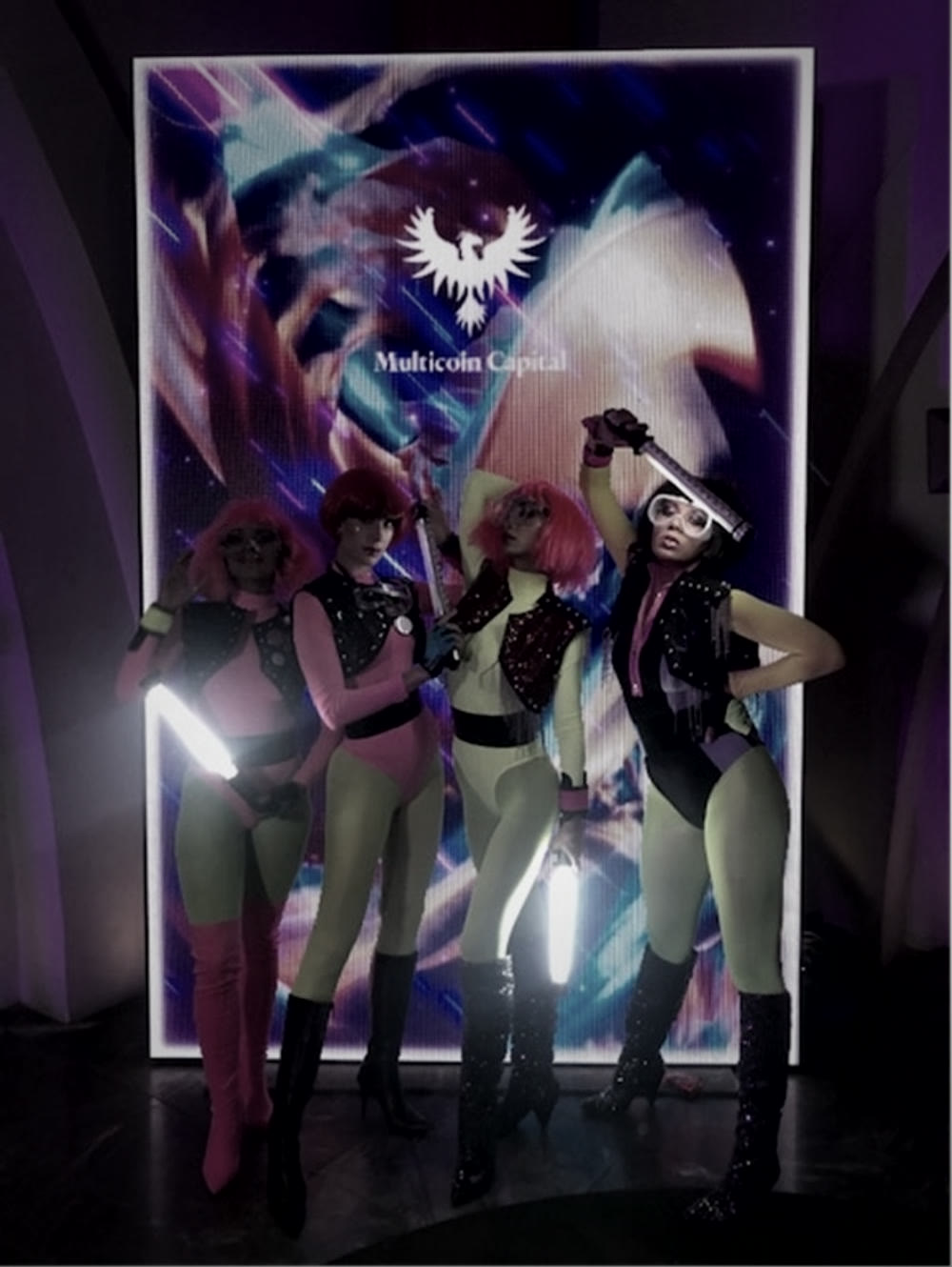 Quatro performers com tubos de luz diante de um painel iluminado