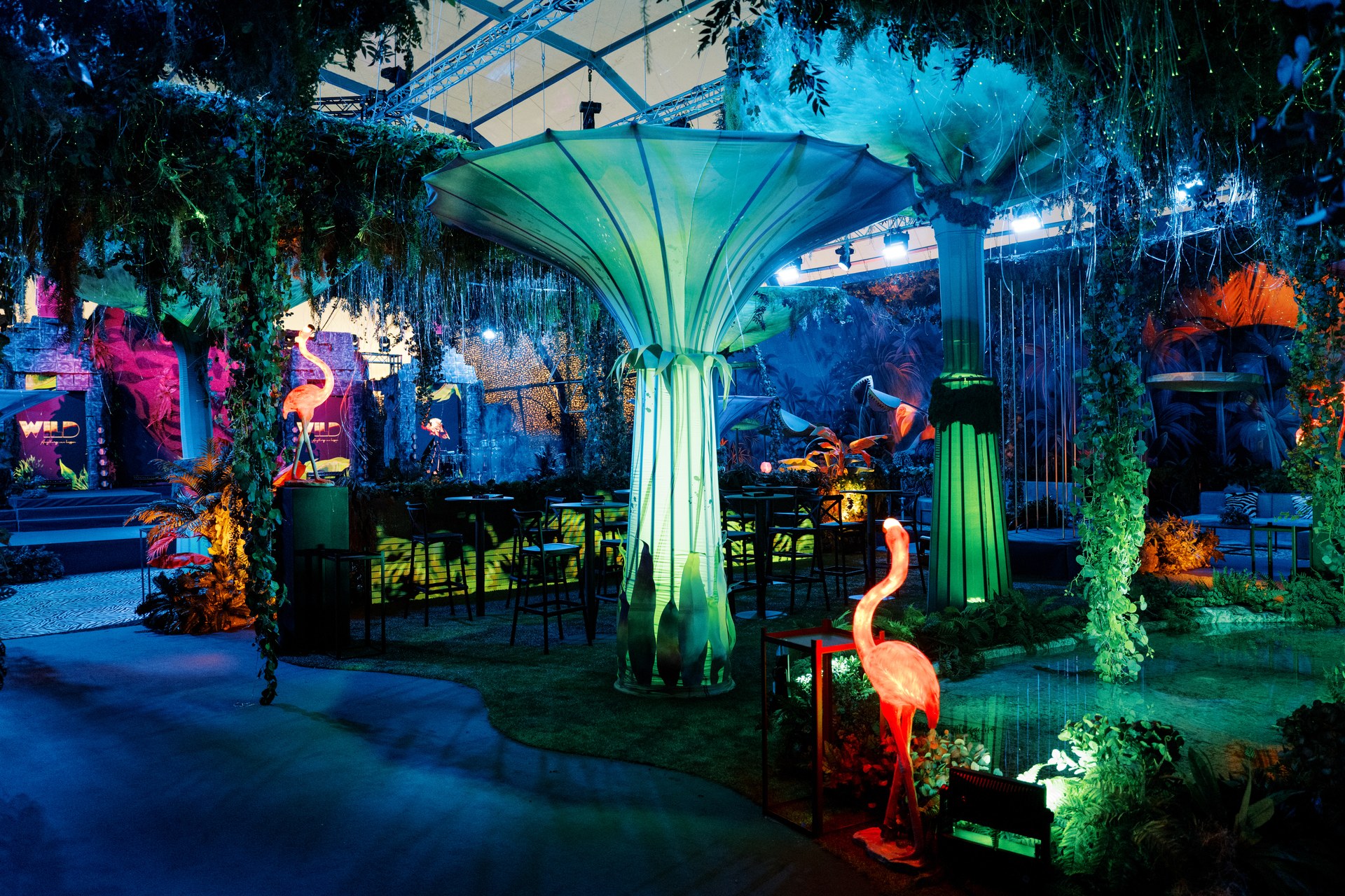 Espaço de evento montado à noite: árvore luminosa em verde e azul, mesas altas e flamingos entre a vegetação