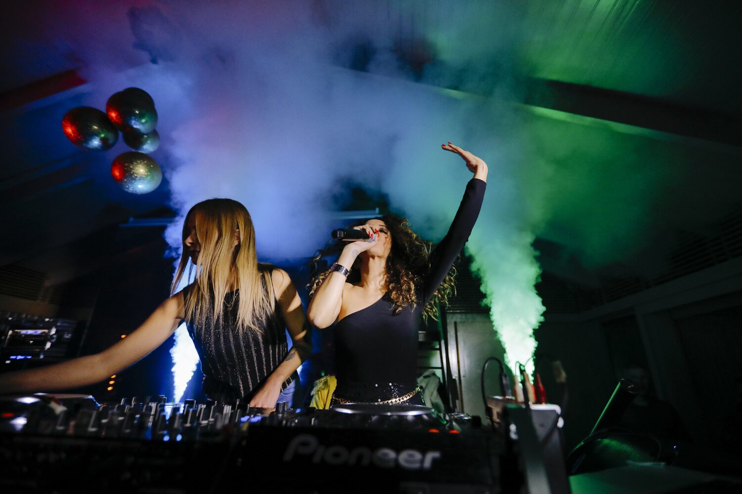 Duas DJs em cabina, entre fumo e luz verde